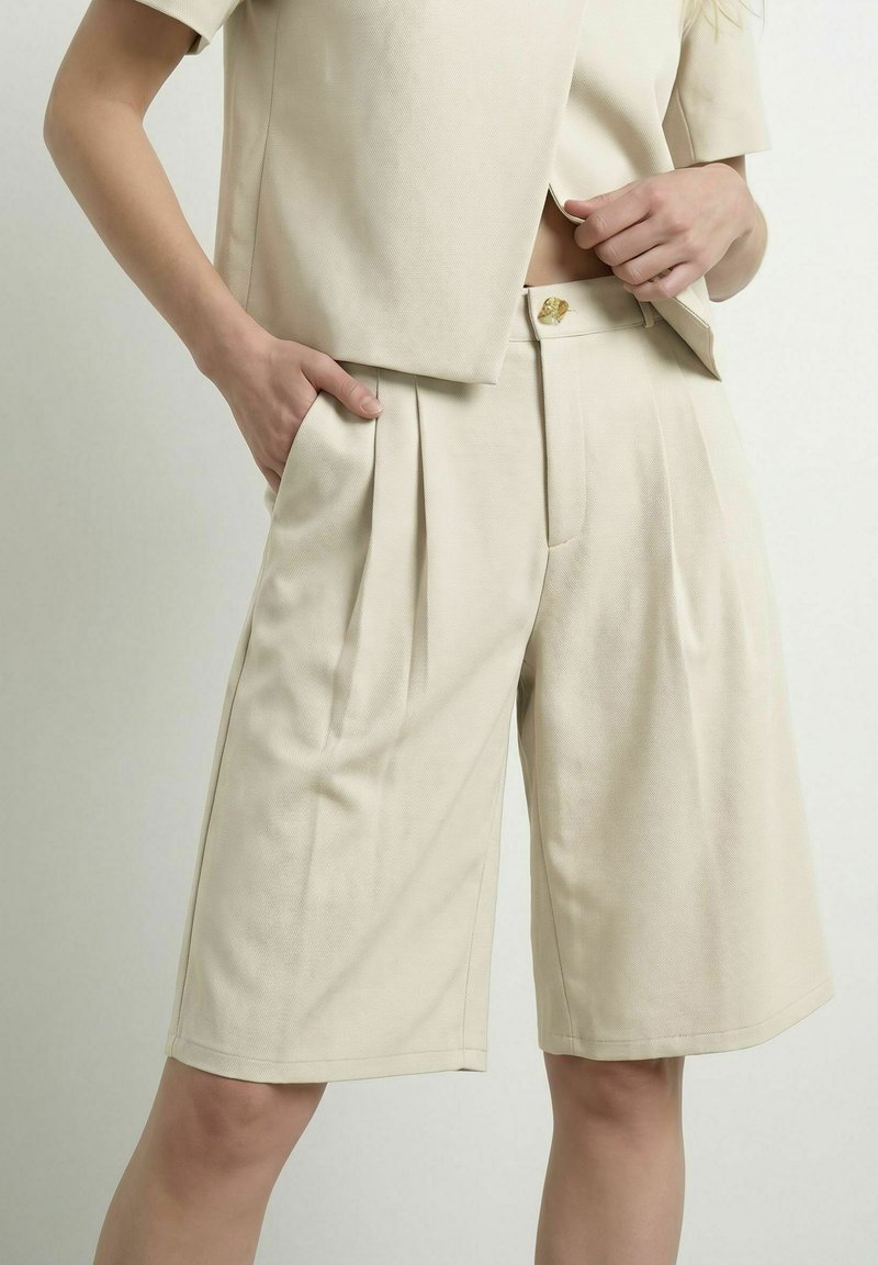 Femme portant un short beige ajusté mi-genou avec des plis et des poches, associé à un blazer à manches courtes assorti, mains posées sur la taille.