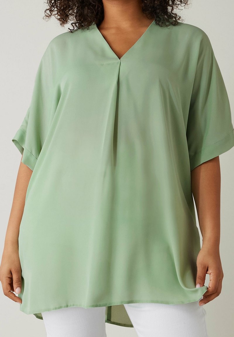 Femme portant un chemisier ample vert clair à manches courtes avec un col en V et un pantalon blanc, tenant l'ourlet du chemisier.