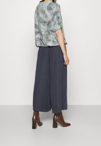 Blusa ampia con un motivo astratto blu e nero abbinata a pantaloni a gamba larga blu navy e stivali con tacco marroni. Texture del tessuto liscia.