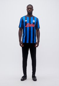 Vyras stovintis, dėvintis juodai mėlynai dryžuotą futbolo marškinėlį, juodas kelnes ir juodus futbolo batelius, stovintis prie paprasto balto fono.