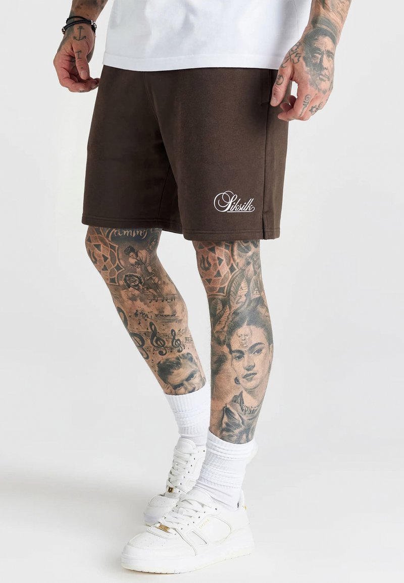 Férfi, sötétbarna rövidnadrágban, rajta "SikSilk" logó, fehér zokni, fehér tornacipő, valamint kiterjedt tetoválások a lábakon és a kezeken.