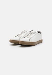 White lace-up shoes with a smooth leather upper, contrast stitching, and a brown rubber sole. Minimalist design with rounded toe shape.