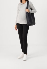 Grijze langemouwen top, zwarte broek en witte sneakers. Een zwarte tote bag met een platte bodem en een verstelbare band wordt in één hand vastgehouden.