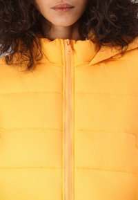 Veste en duvet jaune avec fermeture éclair à l'avant, présentant un design matelassé et une capuche. Texture lisse avec une fermeture éclair contrastante.