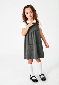 Grau gekleidetes Baumwollkleid mit zwei vorderen Taschen, verstellbaren Trägern und einem gerafften Rock. Kombiniert mit einem weißen Shirt und kniehohen gemusterten Socken.