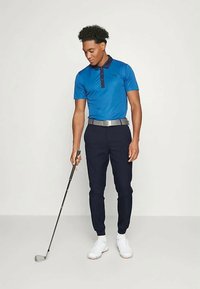 Homem em polo azul e calças azul-marinho, segurando um taco de golfe, olhando para a cabeça do taco, de pé contra um fundo claro e liso.