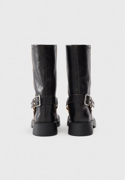 Bottes noires en cuir, montantes jusqu'au genou, avec une finition lisse, une sangle décorative sur le côté, des accessoires en métal et une semelle en caoutchouc épaisse.