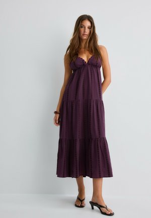 Frau mit langen braunen Haaren, die ein ärmelloses violettes Stufen-Midikleid und schwarze Sandalen mit Absatz vor einem schlichten hellen Hintergrund trägt.