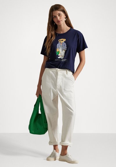 Dark blue cotton t-shirt with a graphic print, white loose trousers, green bag, and light sneakers. Simple design with casual fit.