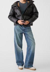 Chaqueta acolchada negra con capucha, con un diseño acolchado. Un par de jeans de pierna ancha en azul claro con un ligero desgaste. Zapatillas blancas con suelas beige.