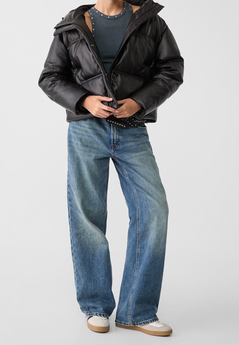 Chaqueta acolchada negra con capucha, con un diseño acolchado. Un par de jeans de pierna ancha en azul claro con un ligero desgaste. Zapatillas blancas con suelas beige.