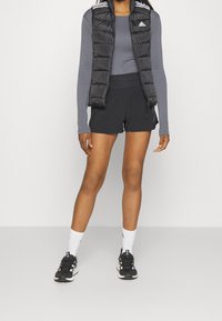 Svart pufferväst med vita ränder på axlarna över en grå långärmad topp, i kombination med svarta shorts och svarta träningsskor.