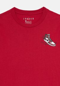 T-shirt en coton rouge avec un graphique d'une chaussure montante ornée d'accents noirs et blancs et de lignes vertes, au design de col rond.