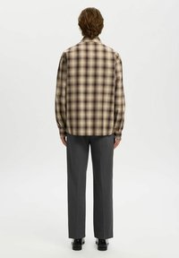 Camisa de cuadros de manga larga en tonos beige y marrón, con un corte relajado y dobladillo redondeado, combinada con pantalones de vestir grises y zapatos negros.