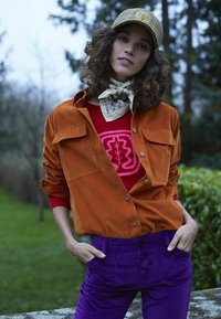 Chemise en daim orange à boutons sur un pull rouge avec un graphisme rose, associée à un pantalon en velours côtelé violet taille haute et une écharpe à motifs.