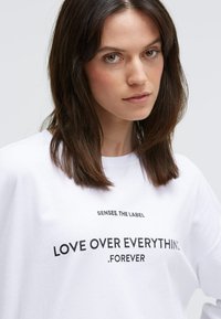 Weißes Baumwoll-T-Shirt mit schwarzem Druck: "SENSES. DAS LABEL LIEBE ÜBER ALLEM . FÜR IMMER." Einfaches Design mit Rundhalsausschnitt und kurzen Ärmeln.