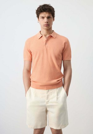 Hombre con una camiseta polo de manga corta color melocotón y shorts de color crema, de pie con las manos en los bolsillos frente a un fondo liso.