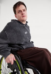 Pelēks kapučs ar logo, apvienots ar vaļīgām brūnām biksēm, ko valkā cilvēks ratiņkrēslā ar zaļiem riteņu akcentiem.