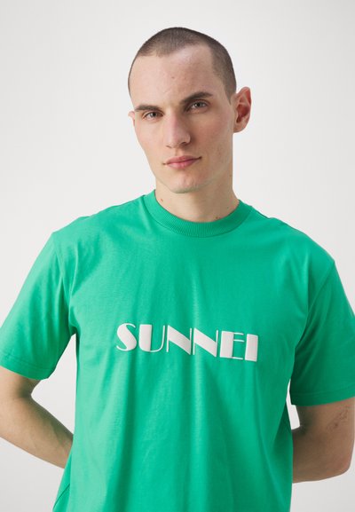 Grønn bomull T-skjorte med en stor hvit "SUNNEI" logo i en stilisert skrifttype. Korte ermer og klassisk rund hals. Glatt tekstur.