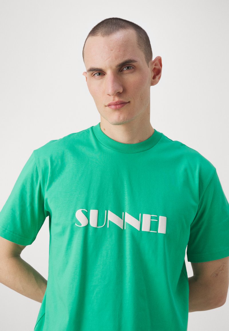T-shirt in cotone verde con un grande logo bianco "SUNNEI" in un font stilizzato. Maniche corte e scollo a giro classico. Texture liscia.