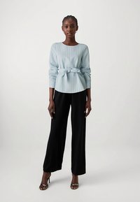 Ανοιχτό μπλε πουκάμισο μακρυμάνικο με κάθετες ρίγες, δεμένο στη μέση, σε συνδυασμό με μαύρο παντελόνι φαρδύ στη μπότα, φορεμένο με μαύρα πέδιλα με λουράκια.