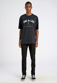 Camiseta negra de manga corta con el texto gráfico blanco "JUNK de LUXE" y logo de corona, combinada con pantalones vaqueros negros y zapatillas en blanco y negro.
