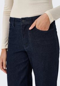 Personne portant un jean bleu foncé avec une main dans la poche avant et un haut beige à manches longues visible jusqu'à la taille.