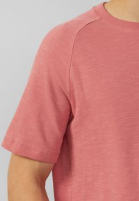 Koszulka z krótkim rękawem, okrągłym dekoltem w jasnoróżowym kolorze, wykonana z miękkiego, teksturowanego materiału z delikatnym melanżowym wzorem oraz widocznymi detalami szycia.