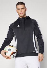 Svart treningshette med hvite detaljer og glidelås, som har Adidas-logoen. Modellen holder en flerfarget fotball. Hvite bukser er synlige.