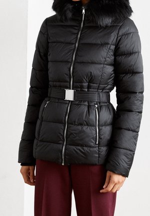 Veste matelassée noire avec fermeture éclair frontale, taille ceinturée, poches zippées et un grand col en fourrure texturée, portée avec un pantalon bordeaux foncé.