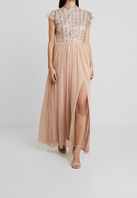 Maxiklänning i nude med genomskinlig paljettkorsett, korta ärmar och en hög slits. Lättviktsmaterial med en flödande kjoldesign.