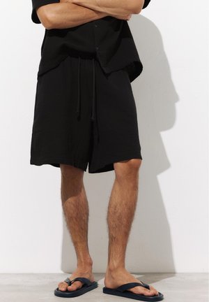 Homme portant une chemise noire boutonnée, un short noir jusqu'aux genoux et des tongs noires, debout, les bras croisés contre un mur blanc.