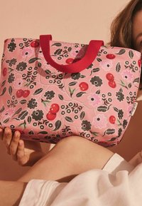 Růžová taška přes rameno s květinovým vzorem, která obsahuje černé a červené třešně, zelené listy a červené uši. Vyrobena z látky s hladkým povrchem.