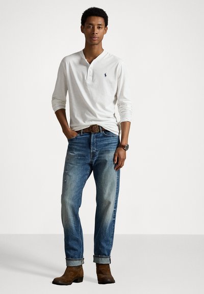 Λευκό χιτώνιο με μακριά μανίκια και μικρό λογότυπο, συνδυασμένο με ξεθωριασμένα μπλε τζιν, καφέ δερμάτινη ζώνη και καφέ μπότες, στέκεται μοντέλο.