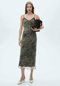 Rochie midi verde măsliniu cu un model ondulat, bretele subțiri și decolteu în V. Modelul ține un obiect maro și poartă pantofi cu vârf ascuțit bej.