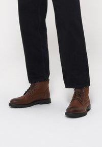 Brązowe skórzane botki za kostkę z sznurowanym wzorem, gumową podeszwą i przeszyciami, w zestawie z czarnymi spodniami.