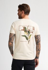 Camiseta de algodón color crema con un estampado botánico en la parte trasera, un logotipo rectangular y texto que dice "NORTHWOODS WISCONSIN U.S.A."