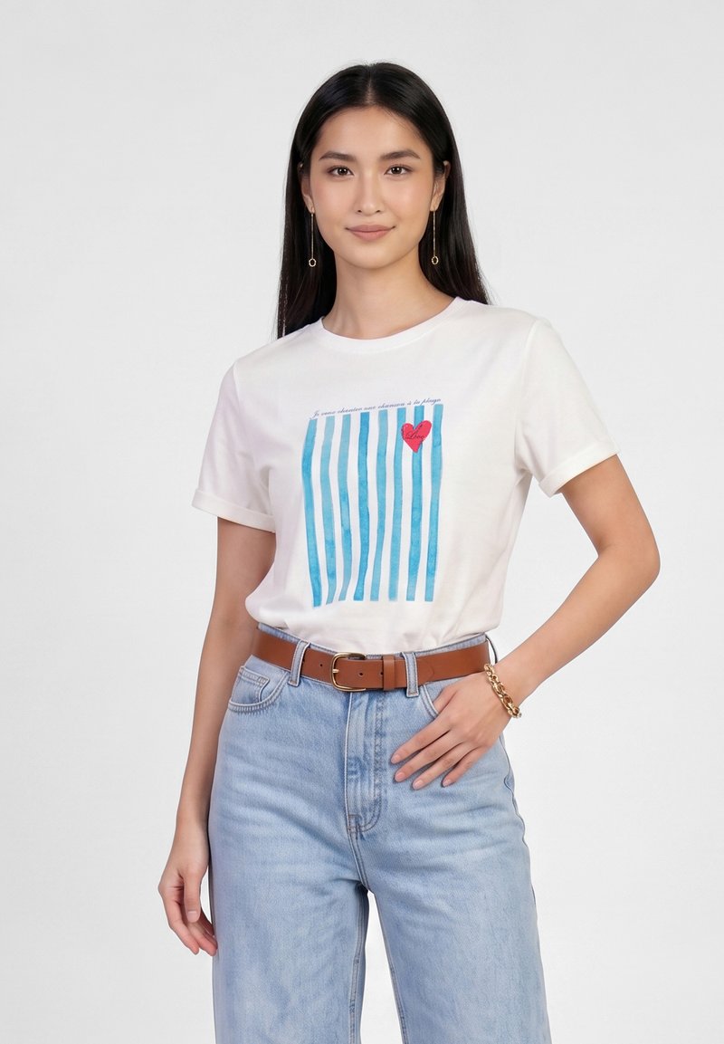 Femme portant un T-shirt blanc avec des rayures verticales bleues et un motif cœur rouge, jean bleu clair, ceinture marron et bracelet en or, posant avec la main sur la hanche.