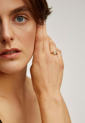 Anillo de oro con un diseño en forma de corazón en el dedo de un modelo, mostrando una textura suave y un acabado pulido sobre un fondo neutro.