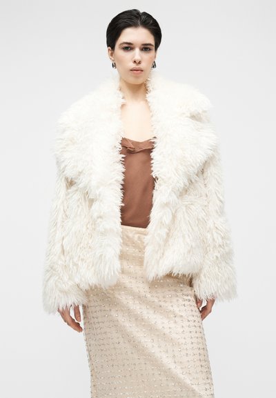 Bela jakna iz umetnega krzna s širokim ovratnikom, ohlapnim krojem in teksturirano tkanino, kombinirana s rjavim topom in bež krilom z bleščicami.