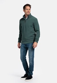 Groene, gestructureerde trui met een ritssluiting bij de kraag, gecombineerd met blauwe jeans en zwarte schoenen. De trui heeft een subtiel verhoogd patroon.