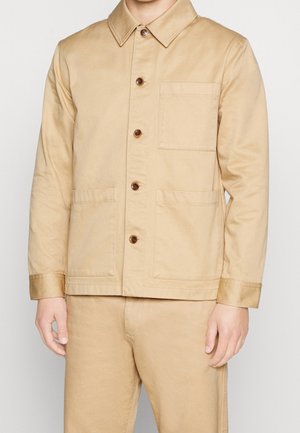 Homme portant une veste beige à boutons avec trois poches avant et un pantalon beige assorti, debout devant un fond uni.