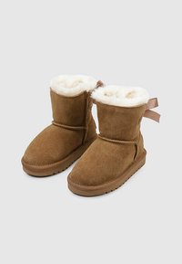 Botas de tobillo de ante marrón con forro esponjoso blanco y detalle de cinta en la parte trasera, exhibidas sobre un fondo gris claro.