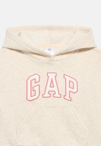 Μωρουδιακό φούτερ με κουκούλα σε κρεμ απόχρωση, με τη λέξη "GAP" σε μεγάλα λευκά γράμματα με ροζ περίγραμμα στο στήθος και εμπρόσθια τσέπη τύπου πουγκί.
