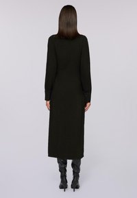 Zwarte lange jurk met rechte snit, hoge halslijn en zijsplitten. Draagt en kniehoge zwarte laarzen. Soepele textuur, minimale details.