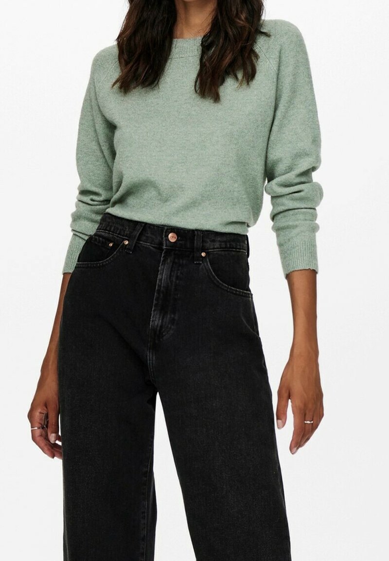 Femme portant un pull tricoté vert clair rentré dans un jean noir ample taille haute, debout devant un fond blanc.