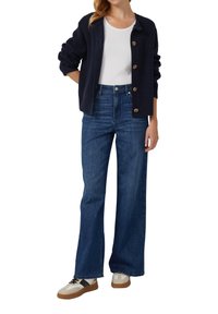 Marineblauwe gebreide cardigan met ronde knopen, witte T-shirt, high-waisted donkerblauwe wijde jeans en beige sneakers met zwarte accenten.