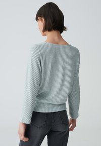 Hellblauer, gerippter Pullover mit weitem Ausschnitt und langen Ärmeln, der einen leicht verkürzten Saum und texturierten Stoff aufweist. Dazu werden schwarze Jeans getragen.