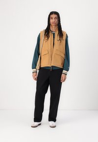 Gilet beige con ampie tasche sopra una felpa verde Tiffany, abbinato a pantaloni neri e sneakers bianche, con un fit rilassato e un design semplice.