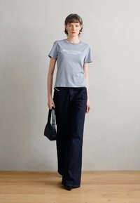 Šedé bavlněné tričko s logem "Marc O'Polo". Kombinováno s tmavě modrými širokými džíny. Modelka drží malou černou kabelku. Pozadí je plochá dřevěná podlaha.